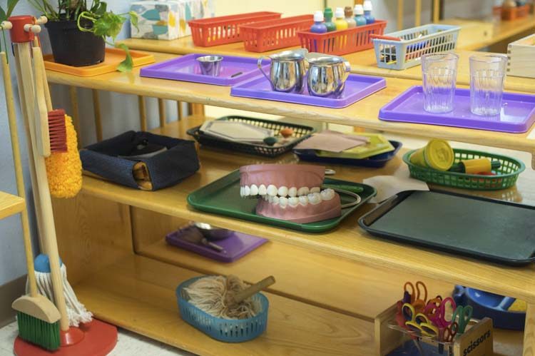 A practical-life shelf set with metal pitchers, glass tumblers, a child-sized broom, and small trays of materials